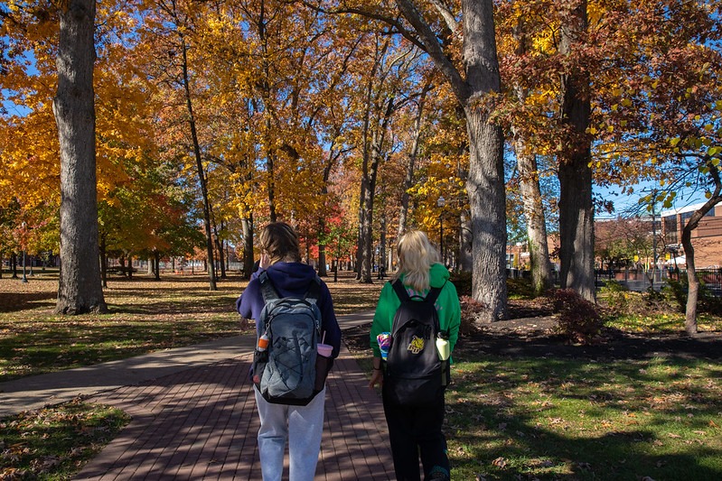 Two students walk through a wooded section of the Rowan University campus during autumn with the leaves changing colors, with their backpacks visible to the camera.
