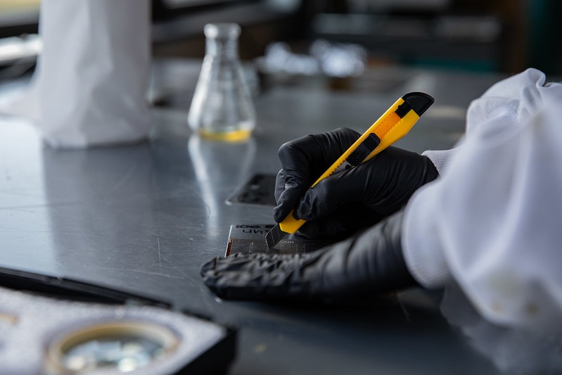 A close up of Rowan University chemical engineering student hands, as they are in black gloves for protection, and they use an X-acto knife to cut something with precision.