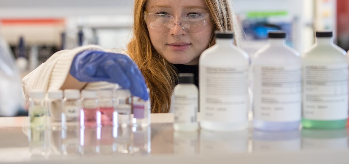 At eye level, a purple gloved hand of a chemical engineering student at Rowan University as she picks up a vial on a rack in a lab.
