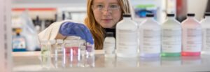 At eye level, a purple gloved hand of a chemical engineering student at Rowan University as she picks up a vial on a rack in a lab.