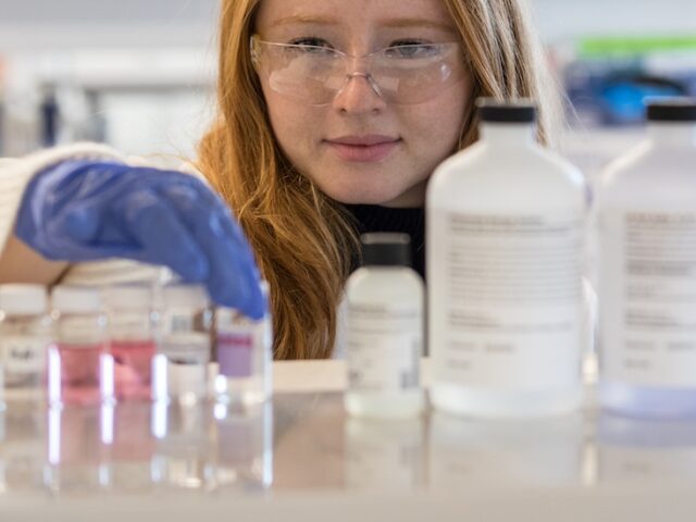 At eye level, a purple gloved hand of a chemical engineering student at Rowan University as she picks up a vial on a rack in a lab.