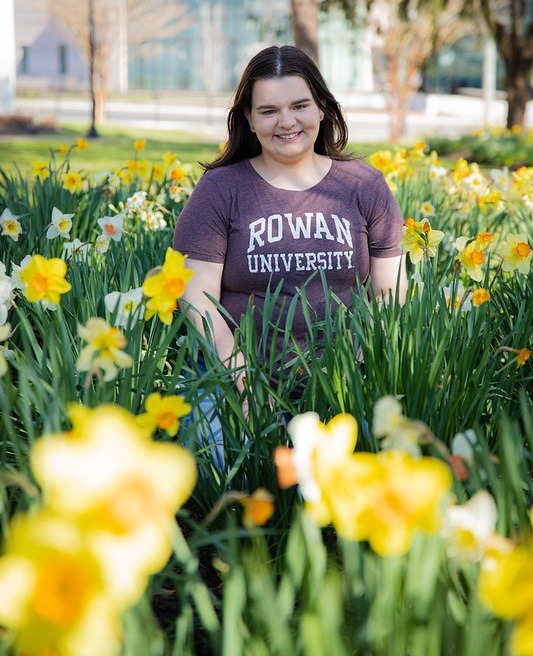 A Rowan University student sits behind a sea of daffodils smiling.