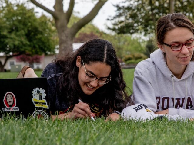 Two Rowan University students lay on their stomachs side by side on Bunce Green, studying with laptops.