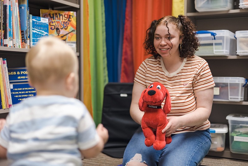 Rowan special education master's student sits in a reading room with a small child, as she holds a red stuffed animal dog.