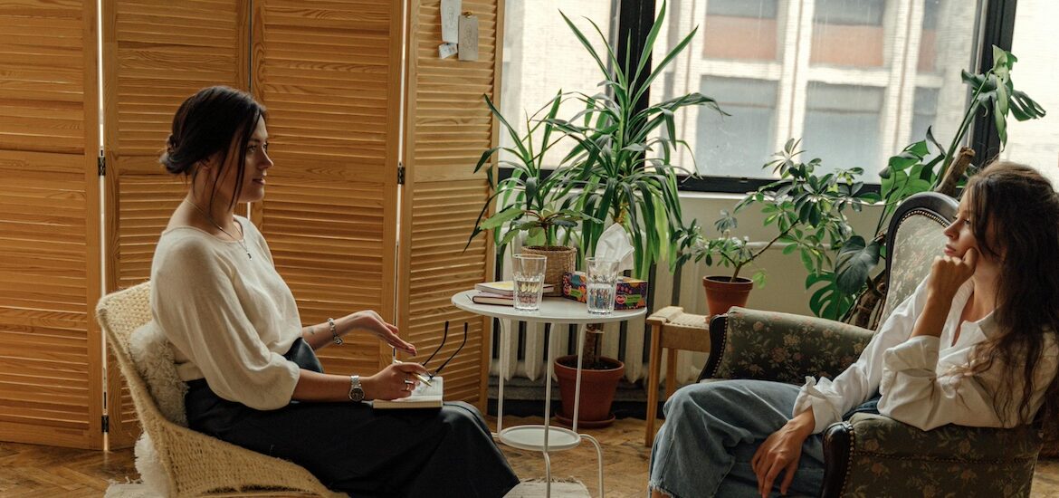 A therapist and a client sit facing each other, with plants around them decorating the office.