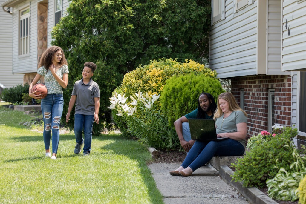 Parents working on a laptop outside their home while two children walk nearby, illustrating how families juggle communication, work, and life—just like students earning a bachelor's in business