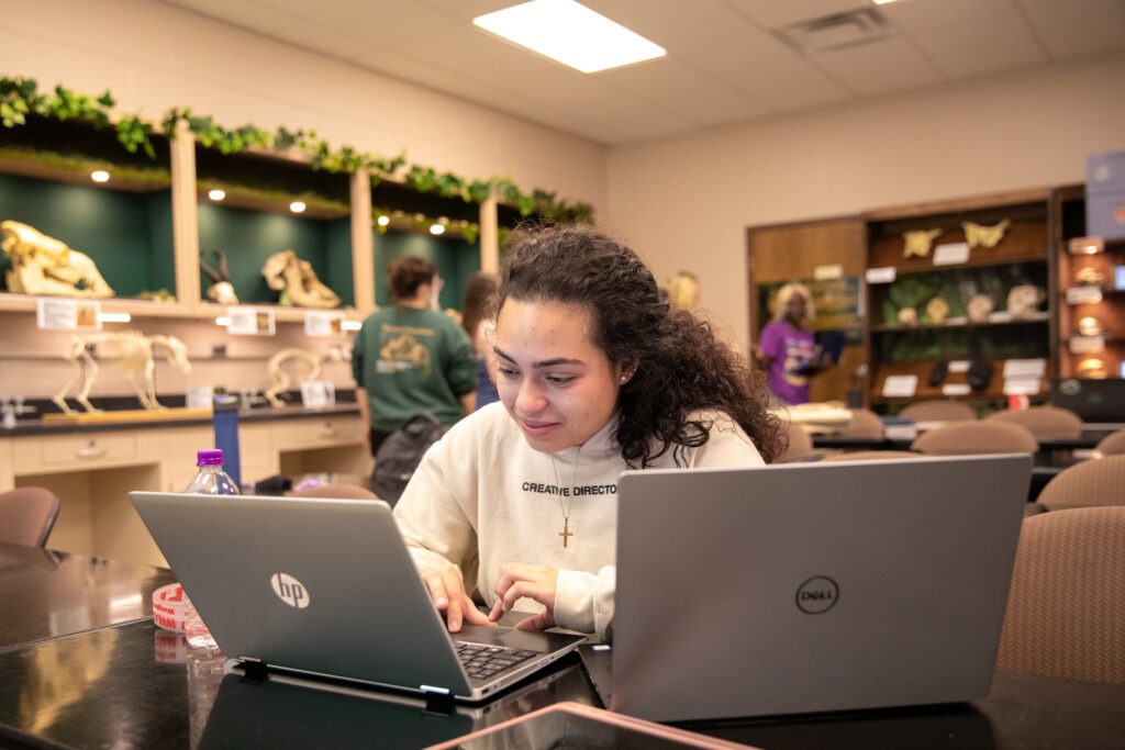 Student using dual laptops in a science lab to complete a tech-integrated assignment, representing skills built in a master’s in educational technology online program