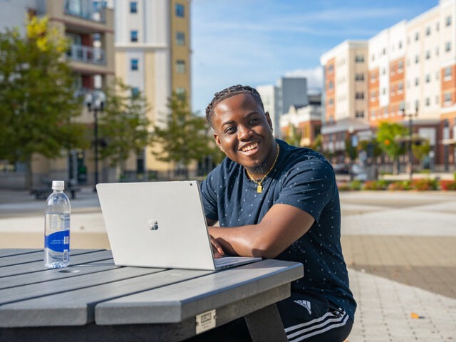 College student smiling while working on a laptop outdoors, representing the flexibility and confidence that come with pursuing an online business bachelor's degree