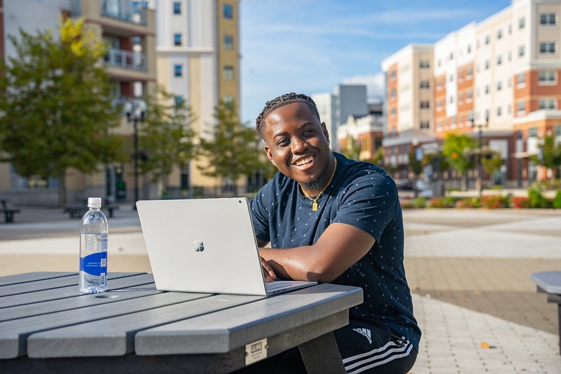 College student smiling while working on a laptop outdoors, representing the flexibility and confidence that come with pursuing an online business bachelor's degree