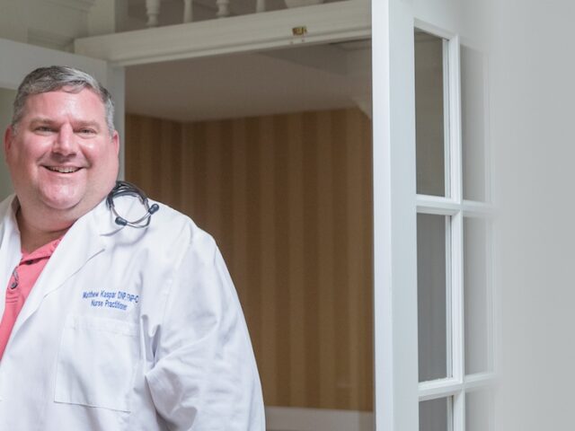 An aspiring CNO Rowan University student enrolled in the master of science of nursing, nurse executive track, smiles at the camera while at work in a hospital wearing a white coat and stethoscope.