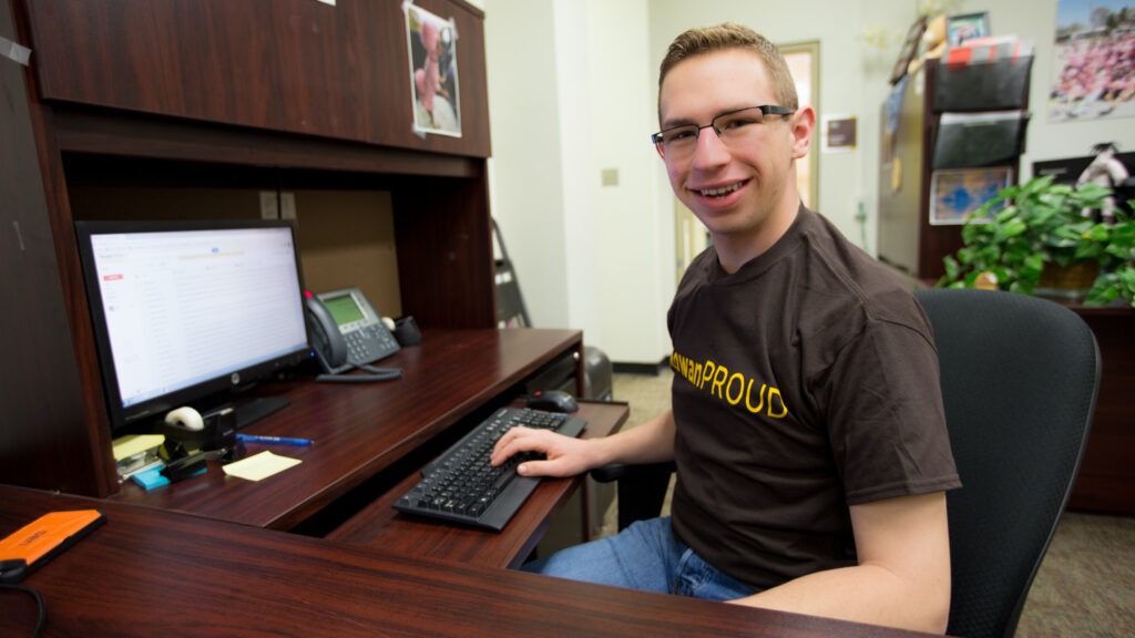 Student sitting down at computer completing online coursework for liberal studies degree, wearing RowanProud shirt
