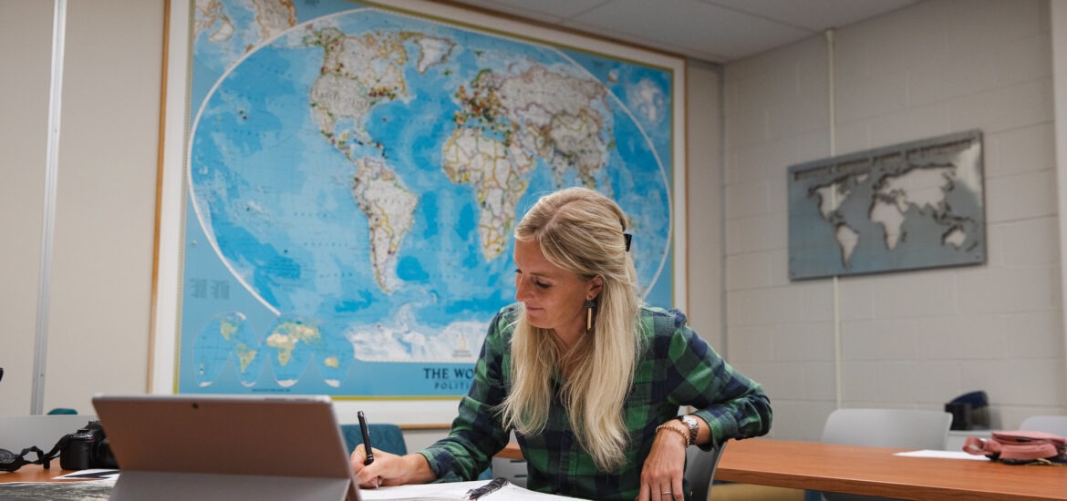 Student completing liberal studies degree coursework online sitting down at table with map on wall