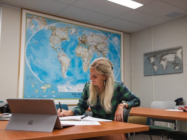 Student completing liberal studies degree coursework online sitting down at table with map on wall