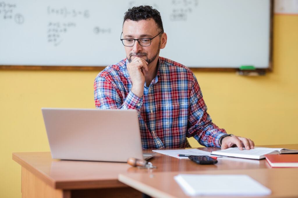 Teacher in classroom sitting down to complete online coursework for master's in educational technology program.