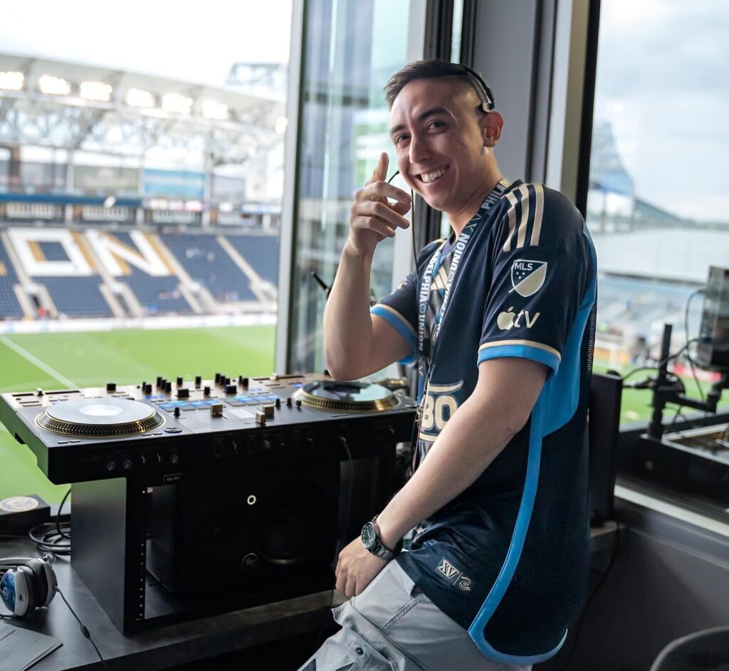 JT sits at a DJ turntable, turned away to look at the camera smiling. In the distance in front of him is the Philadelphia Union stadium seating and field. 