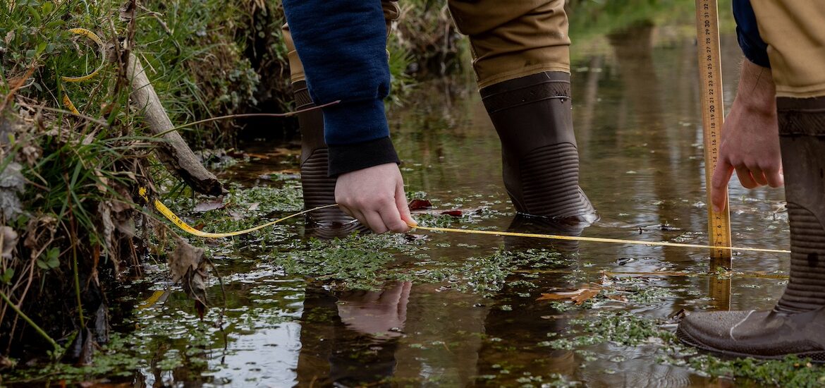 A close up of creek water with environmental science majors' feet and hands working in the water.