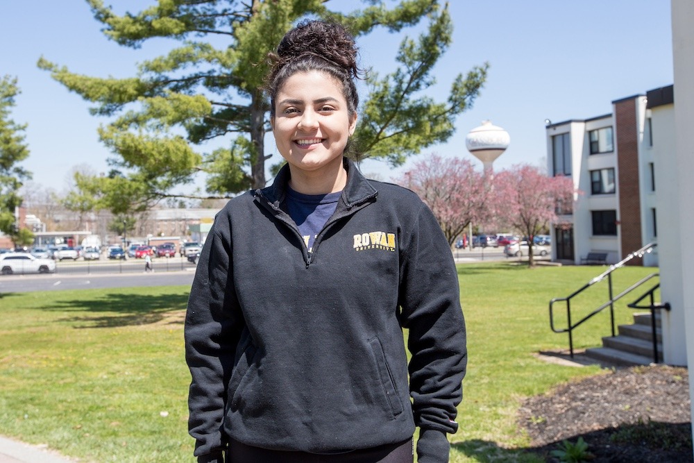 A student stands outside a dorm on a bright day, wering a long sleeved pullover and with her hands in her pockets. 