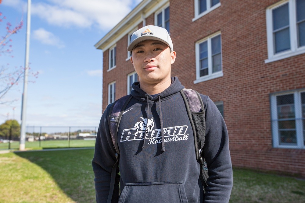Wearing a Rowan Racquetbll hoodie and a baseball cap, a student stands for a portrait outside of Willow Hall. 