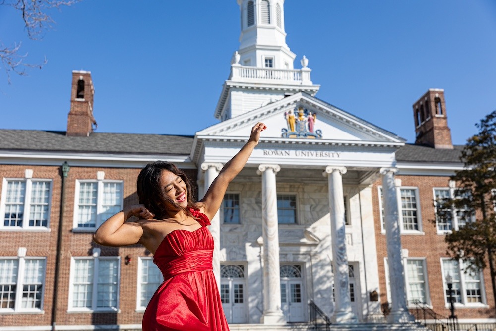 A Rowan student wearing a red dress flexes her arm in the air to show excitement, with a big smile on her face and the iconic marble columns of Bunce Hall behind her.