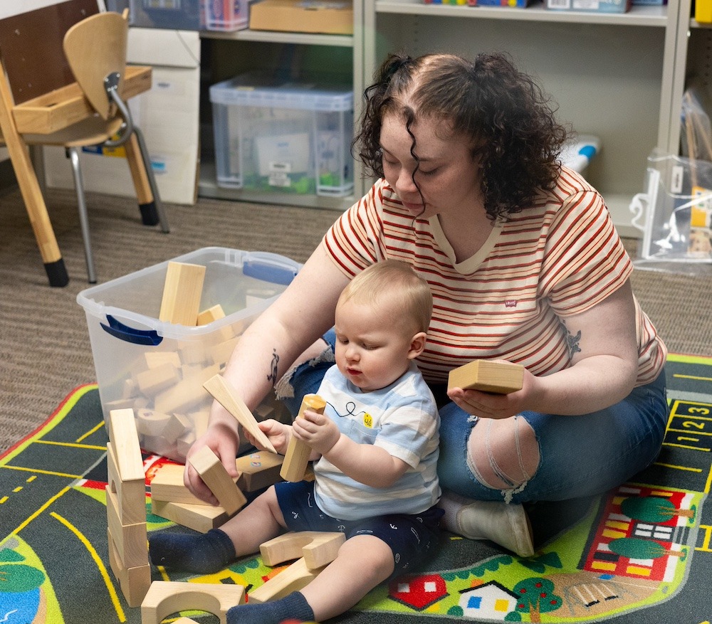 A student sits on the ground with a one year old who is playing with blocks.