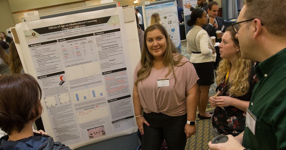 A student presents her research at a poster session, with the poster on display by her side and three people around her reading it and learning.