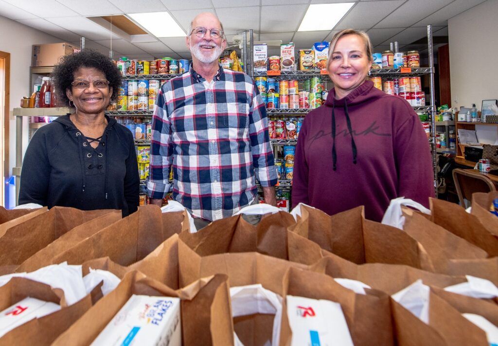 Three people stand side by side at a food bank, smiling at the camera, with rows of brown paper bags in front of them and rows of shelving with canned foods behind them.