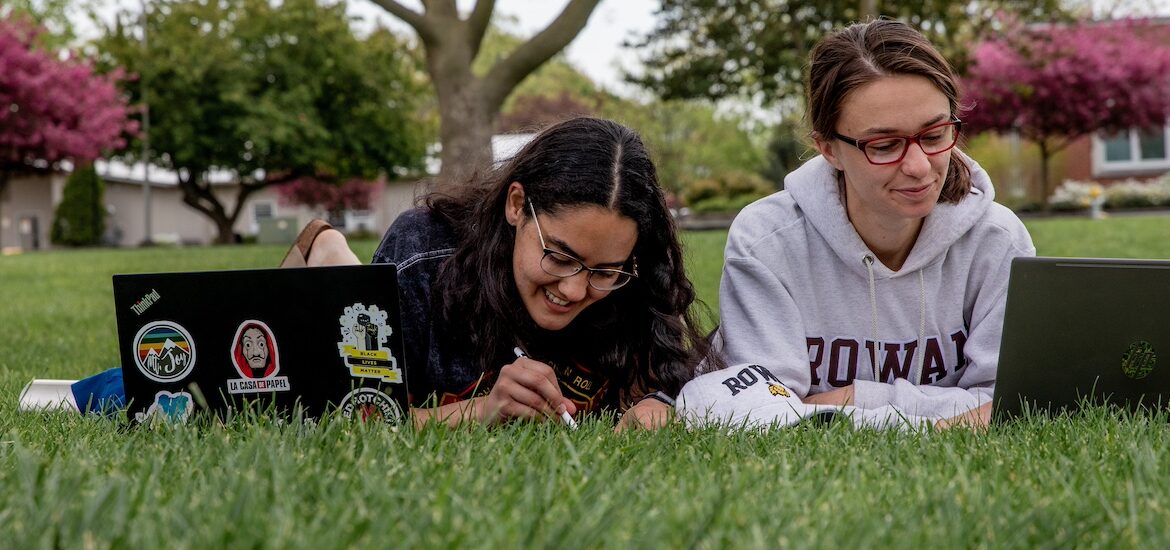 Two students study side by side laying on their stomachs on the lawn, with laptops in front of them.