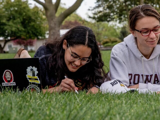 Two students study side by side laying on their stomachs on the lawn, with laptops in front of them.