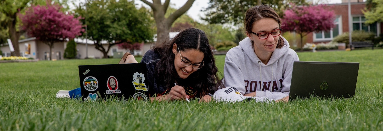 Two students study side by side laying on their stomachs on the lawn, with laptops in front of them.