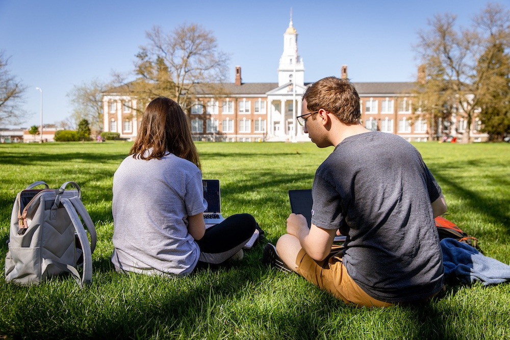 The backs of two students as they study sitting on the grass side by side, witch iconic Bunce Hall ahead of them in the distance.