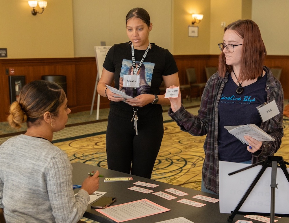 Two law and justice students show a card that says "you can donate plasma" to a person manning a table at the prisoner re-entry simulation.