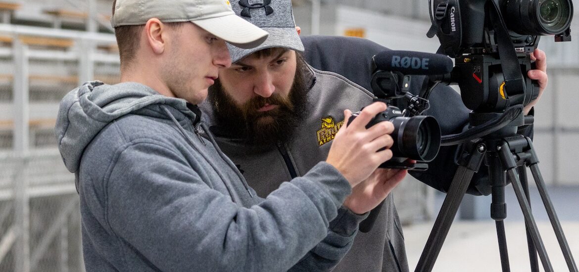 A Rowan radio tv film major works with a supervisor in the field looking at a camera.