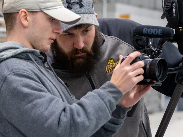 A Rowan radio tv film major works with a supervisor in the field looking at a camera.