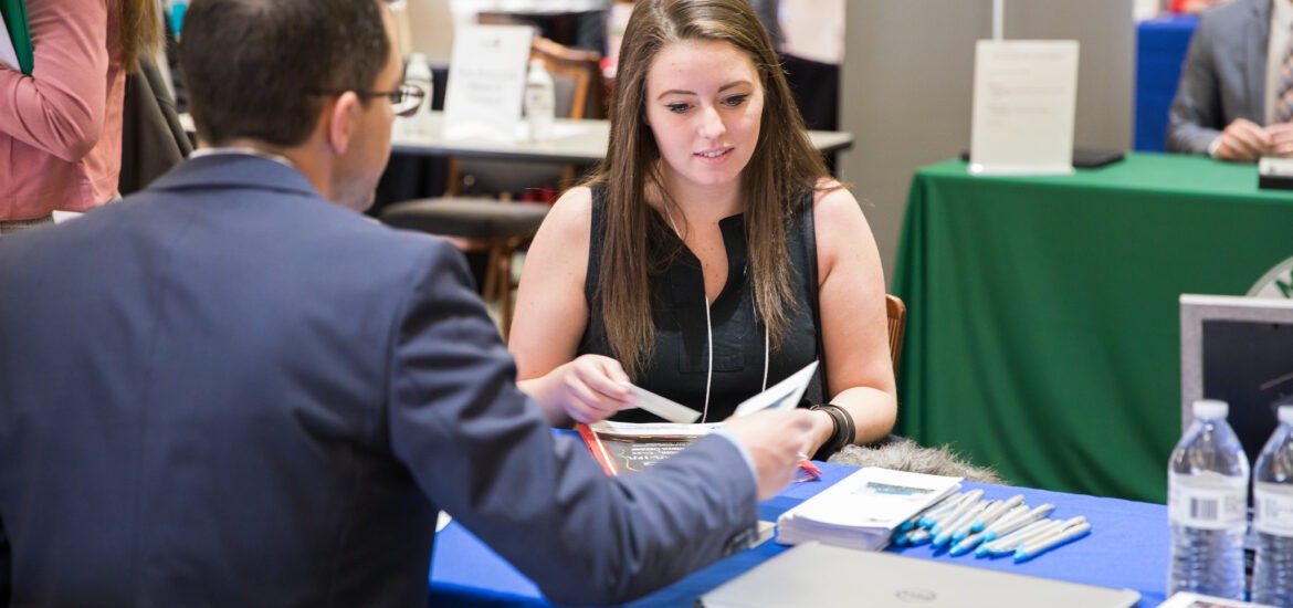 Online MBA student speaking with a recruiter at a career fair table, highlighting how networking helps market your MBA degree.