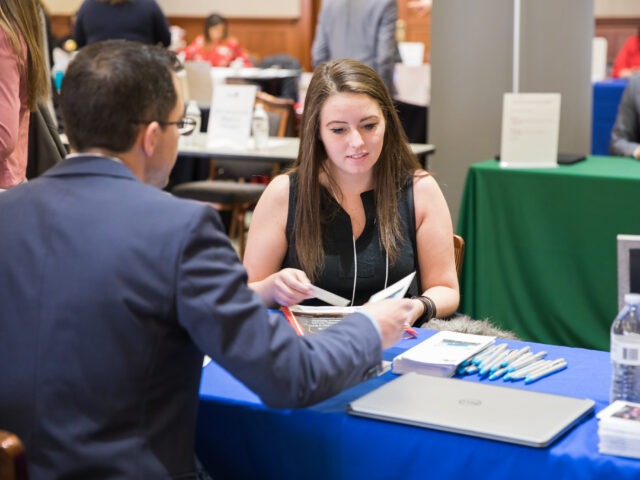 Online MBA student speaking with a recruiter at a career fair table, highlighting how networking helps market your MBA degree.