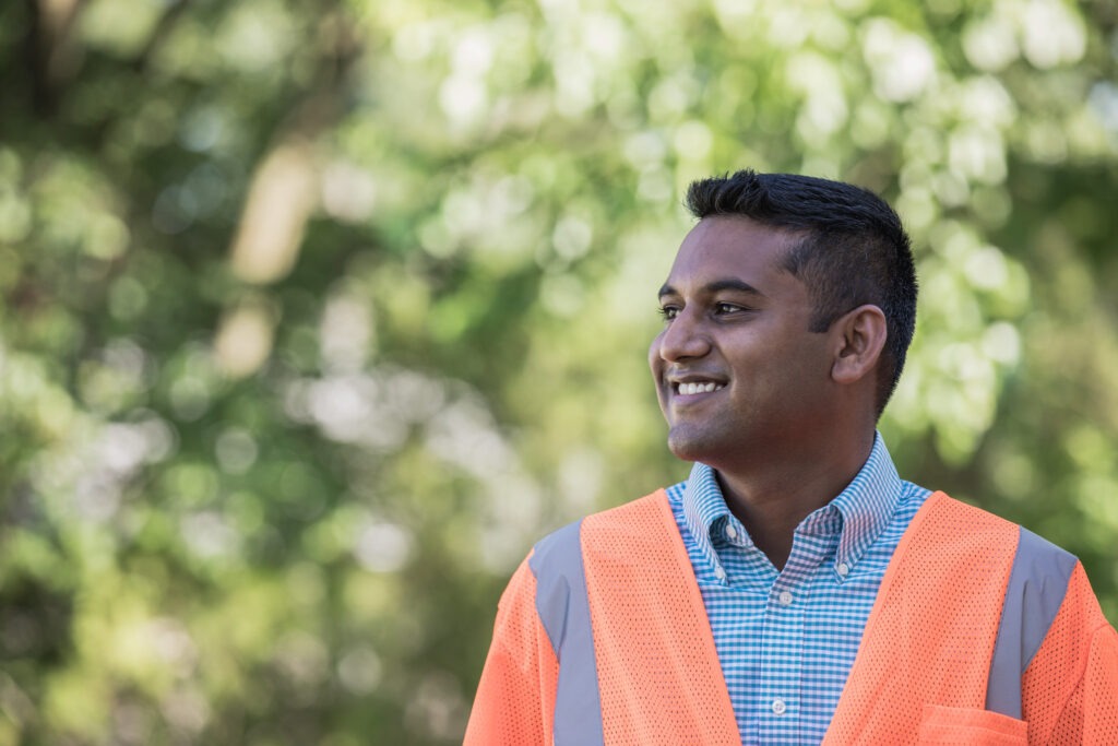 Construction management professional wearing an orange safety vest, smiling outdoors on a sunny day.