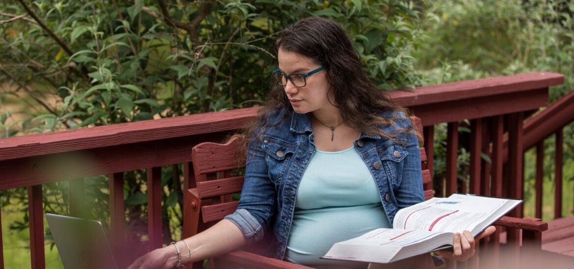 Adult student studying online outdoors with laptop and textbook, balancing education and family life.