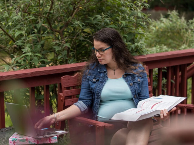 Adult student studying online outdoors with laptop and textbook, balancing education and family life.