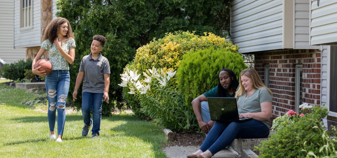 Working parents completing online coursework from home while kids play outside.