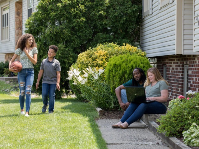 Working parents completing online coursework from home while kids play outside.