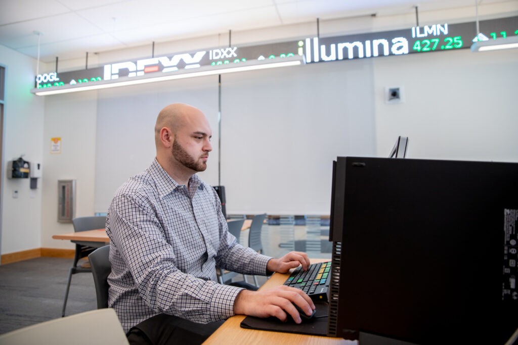 Business professional working at a computer under a stock ticker display, symbolizing financial management career opportunities with an MBA.