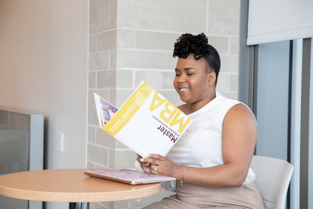 Woman smiling while reading Rowan University MBA folder at a table, symbolizing how to market yourself with an online MBA degree.