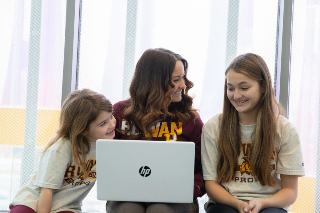 Adult student and children wearing Rowan University shirts studying together on a laptop.