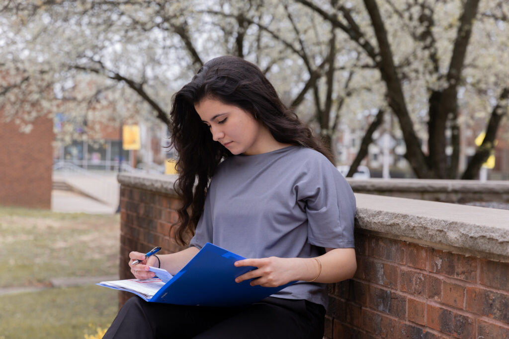 Female student sitting outdoors, writing in a folder and preparing career notes to showcase her online MBA skills.