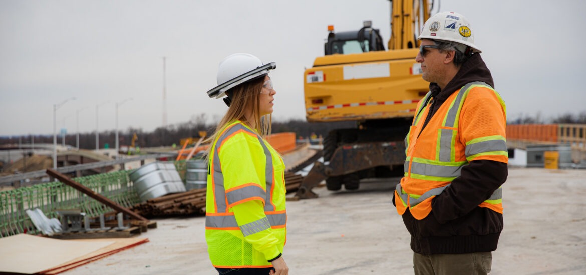 Female construction manager in a yellow safety jacket talking with a male colleague on an active construction site.