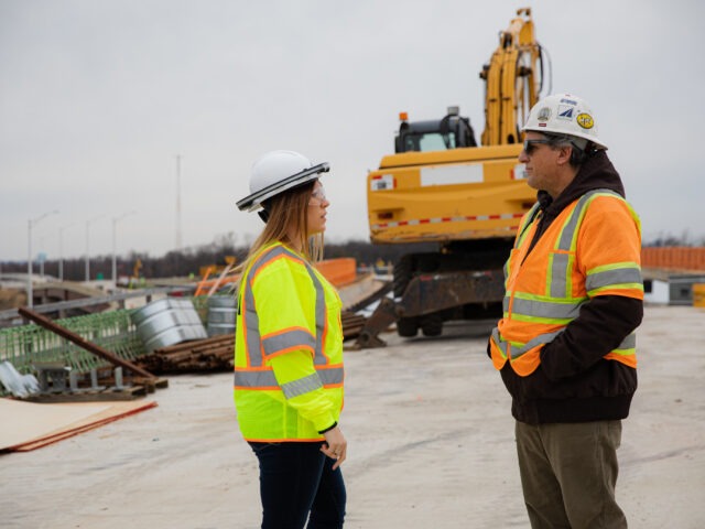 Female construction manager in a yellow safety jacket talking with a male colleague on an active construction site.