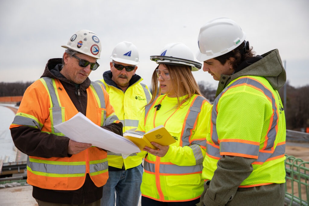 Construction managers reviewing blueprints on a job site wearing safety vests and hard hats.