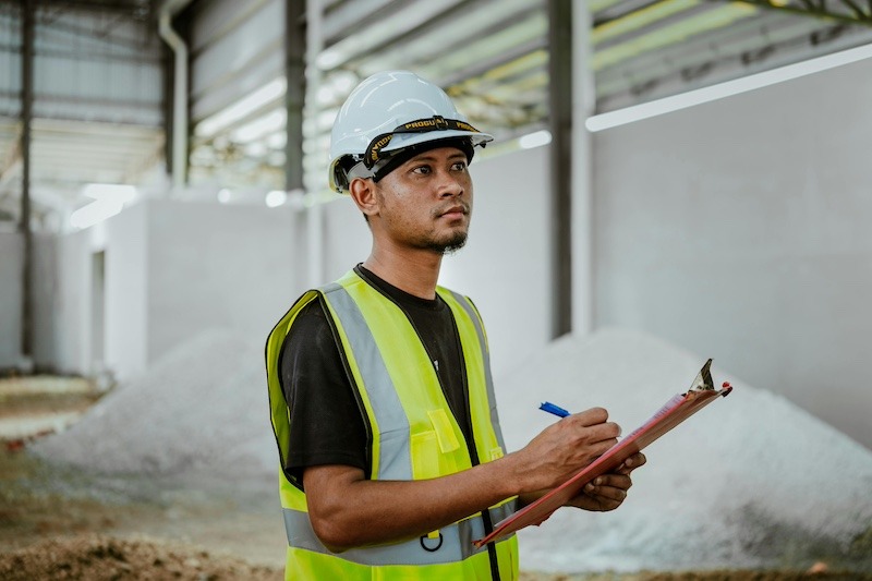 A construction manager holds a clip board, wearing a vest and a hard hat on a job site.