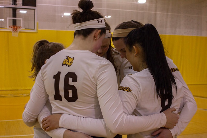 Five women's volleyball players huddle tight on the court, arms around one another.
