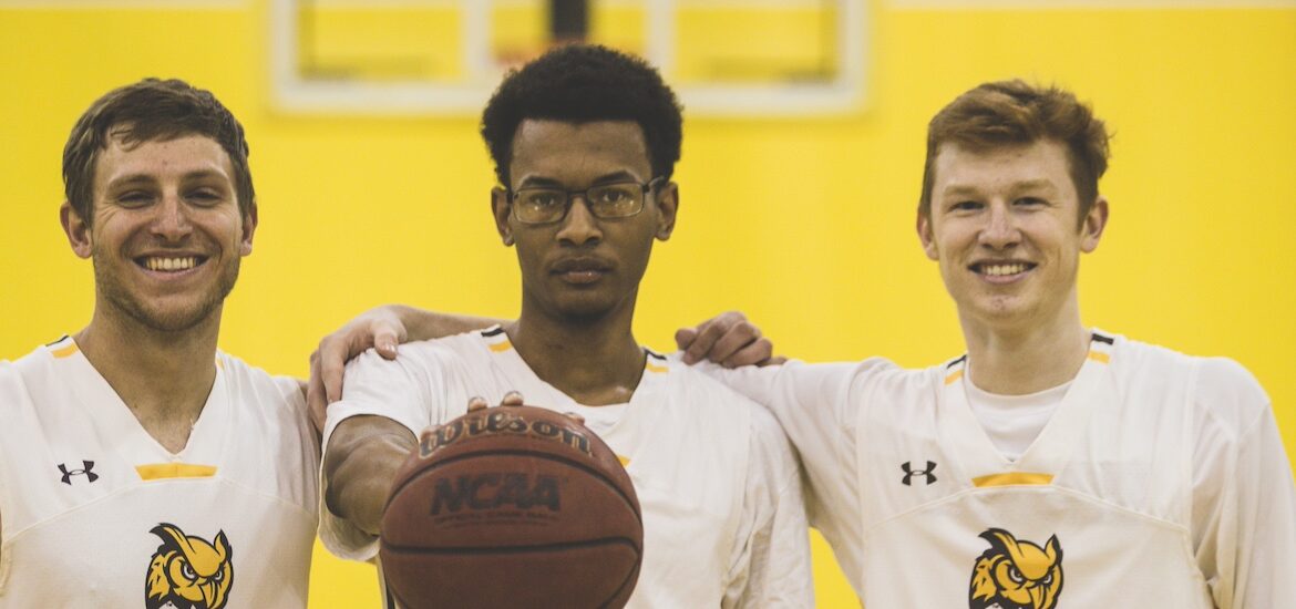Three men's basketball players stand shoulder to shoulder looking at the camera, with the player in the middle palming a basketball and holding it out in front of him.